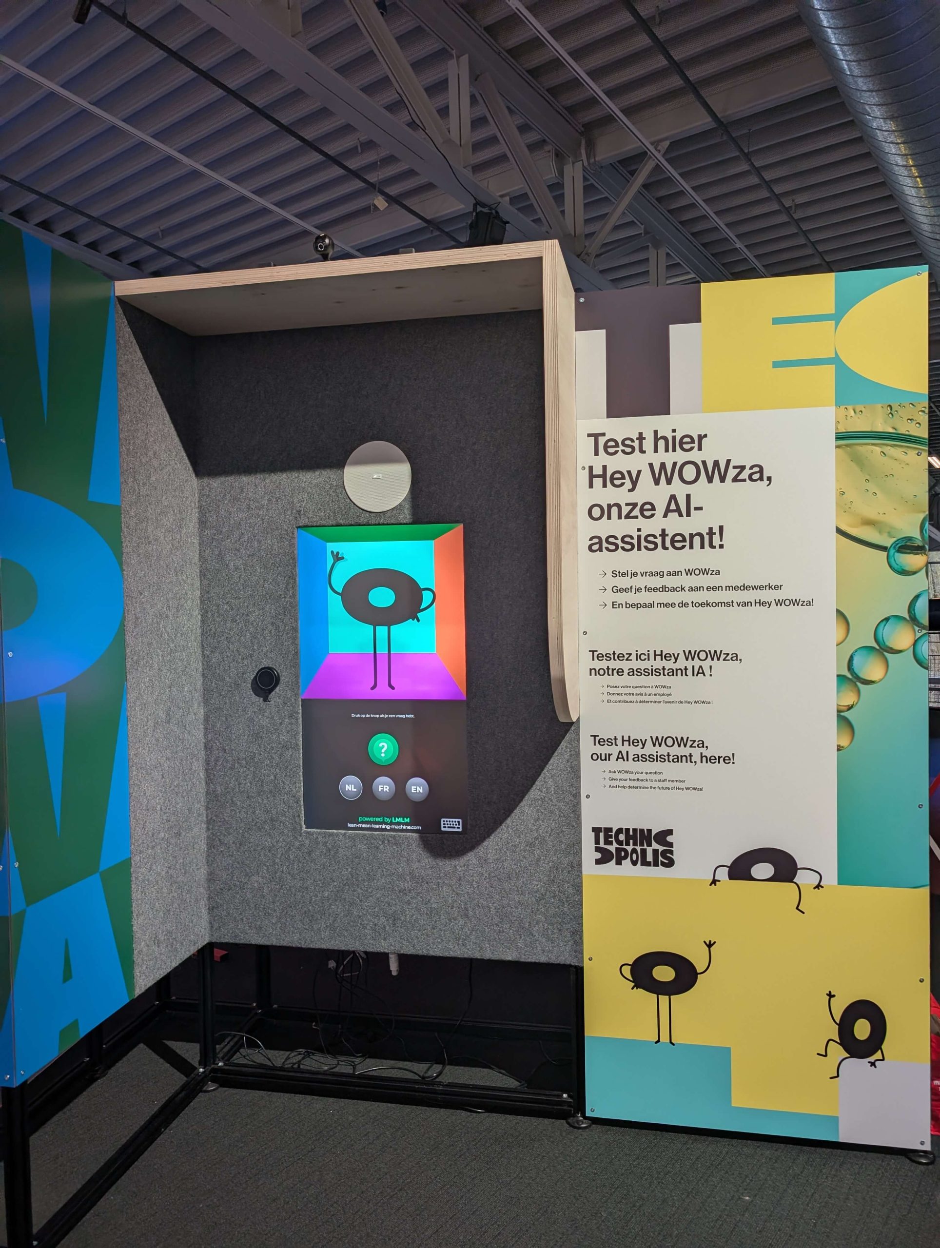 The heyWowza booth at Technopolis: a felt-lined kiosk with Wowza on screen, a green question button, NL, FR and EN buttons, and the Test hier Hey WOWza side panel